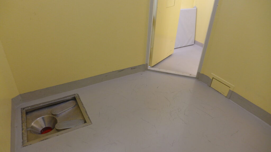Innsiden av celle med gule vegger og toaletthull i gulvet.